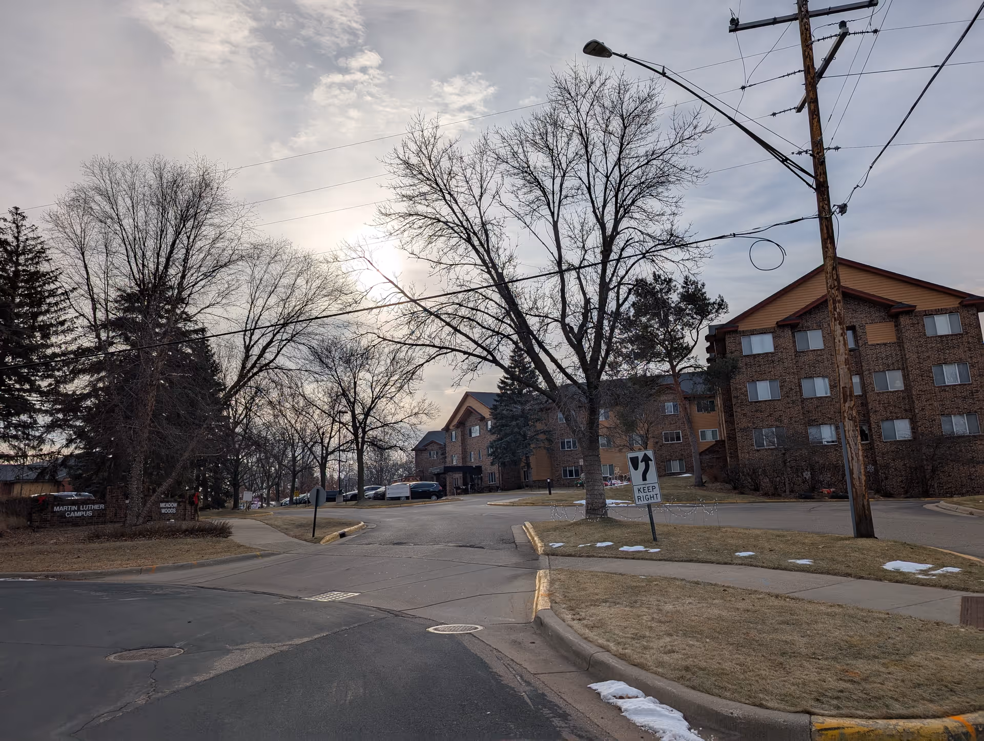 View of the Martin Luther Campus senior living facility exterior with a paved road, leafless trees, and a multi-story brick building under a cloudy sky during late winter or early spring.