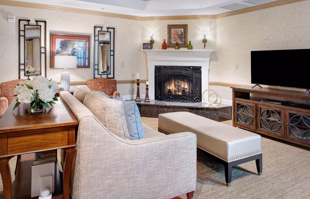 A cozy living room with a beige sofa and blue cushions facing a fireplace with a decorative black screen. There is a long beige bench in front of the sofa, a wooden TV stand with a flat-screen TV, and a wooden side table with a white flower arrangement and a lamp. The walls are light-colored with two vertical mirrors and a framed picture above the sofa. The fireplace mantel has decorative items and two wall sconces with lights.