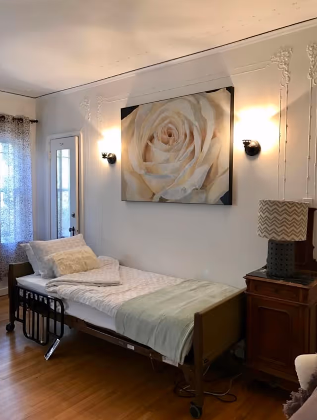 A single bed with white bedding and pillows in a room with wooden flooring. Above the bed is a large painting of a white rose. On the right side of the bed is a wooden nightstand with a lamp featuring a chevron-patterned shade. There is a window with sheer curtains on the left side of the room and a door next to it.