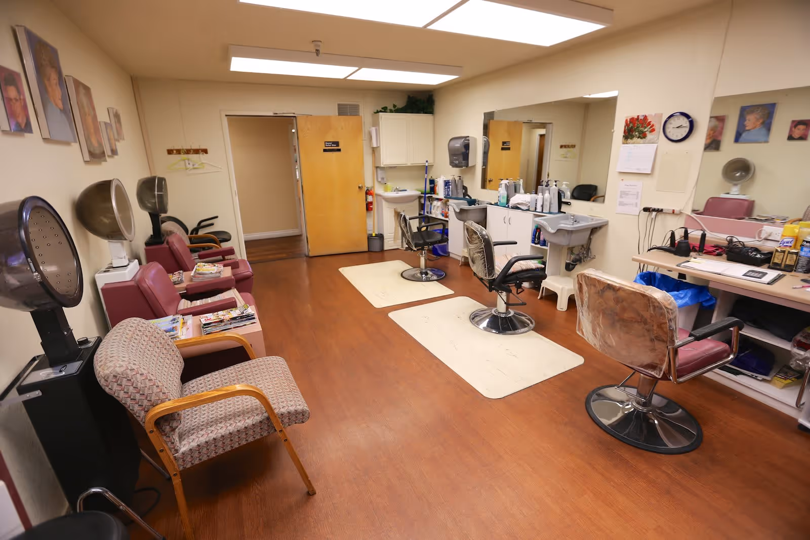 Interior view of a senior living facility's hair salon with several hair drying chairs along the left wall, salon chairs in front of mirrors on the right, and various hair care products on counters. The room has wood flooring, fluorescent ceiling lights, and portraits on the walls.