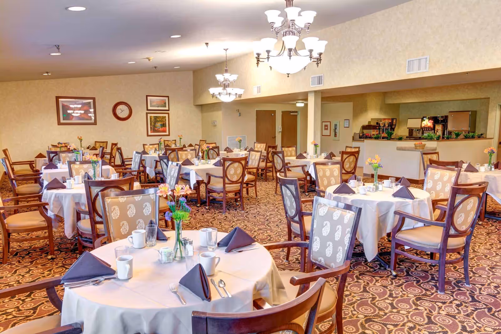 A spacious dining room with multiple round tables covered with white tablecloths, each set with cups, glasses, silverware, and folded dark napkins. The chairs have wooden frames with patterned upholstery. The room features patterned carpet, beige walls with framed artwork and a clock, and multiple chandeliers providing warm lighting. There is a serving area in the background with plants and a basket of fruit.