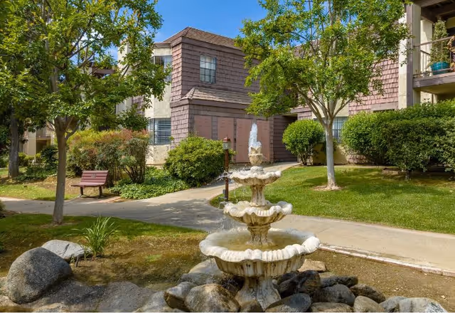 Outdoor area of Oaks of Pasadena featuring a three-tiered stone water fountain surrounded by rocks, green grass, trees, bushes, a bench, and a pathway leading to residential buildings with brown shingle siding under a clear blue sky.
