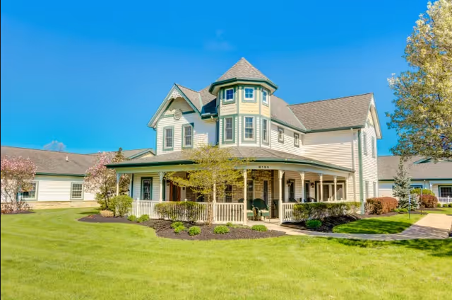 A large, two-story house-style building with a turret and wrap-around porch, surrounded by well-maintained green lawns, shrubs, and trees under a clear blue sky.