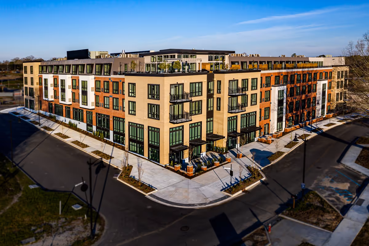 Aerial view of a modern multi-story senior living facility building with large windows, balconies, and a rooftop terrace, surrounded by paved roads and sidewalks under a clear blue sky.