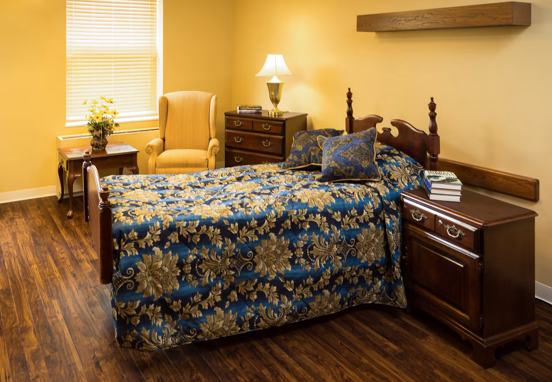 A cozy bedroom with a wooden bed covered in a blue and gold floral bedspread, matching pillows, a wooden nightstand with books, a wooden dresser with a lamp and books, a yellow armchair, a small table with a vase of yellow flowers, and a window with blinds letting in natural light.