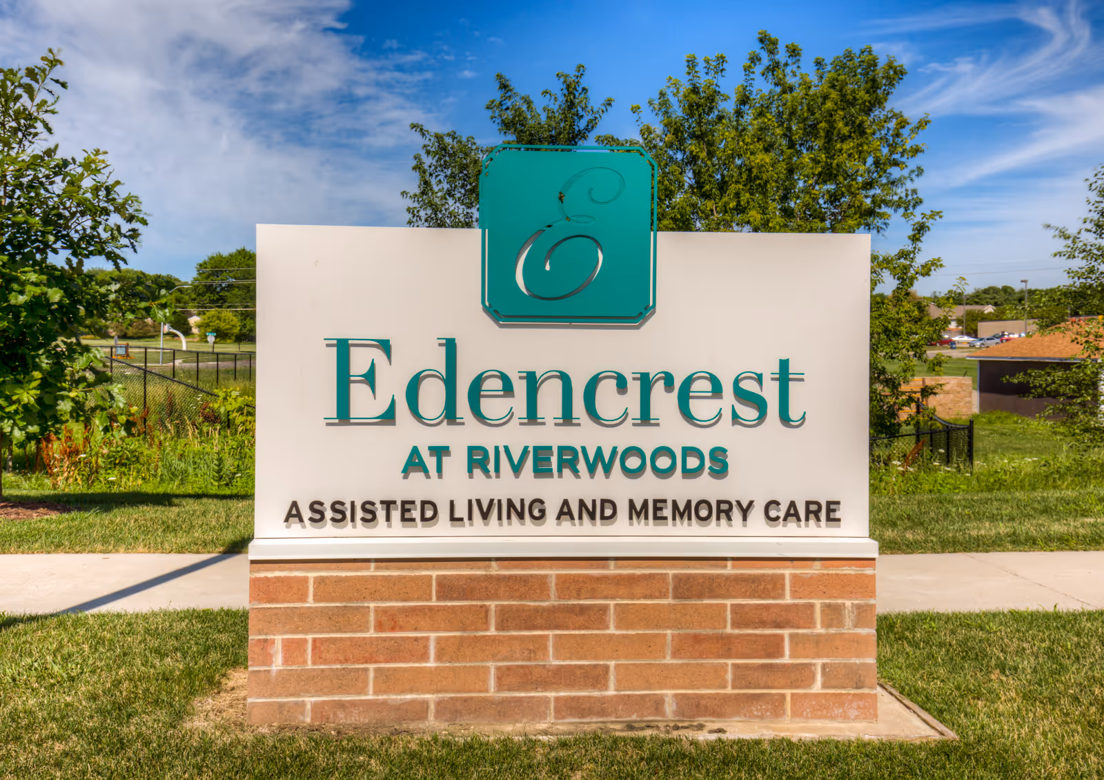 A large brick-and-panel outdoor sign reading "Edencrest at Riverwoods Assisted Living and Memory Care" on a grassy lawn with trees and blue sky behind it.
