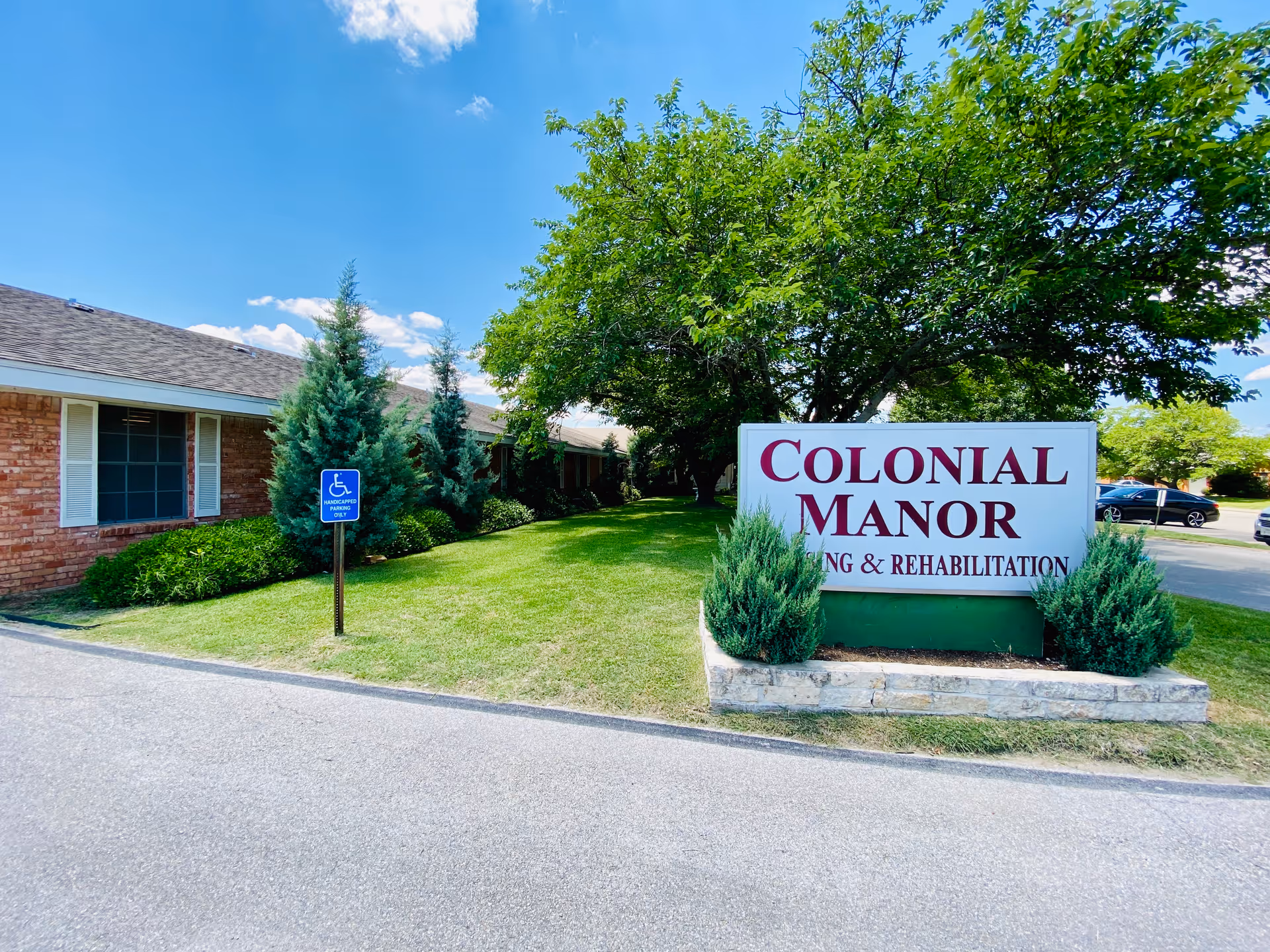 Exterior view of Colonial Manor Nursing & Rehabilitation facility showing a brick building with windows, a green lawn with trees and shrubs, a handicapped parking sign, and a large sign with the facility's name.
