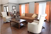 A living room with a brown sofa, two white armchairs, a wooden coffee table, and large windows with orange curtains allowing natural light to fill the space.