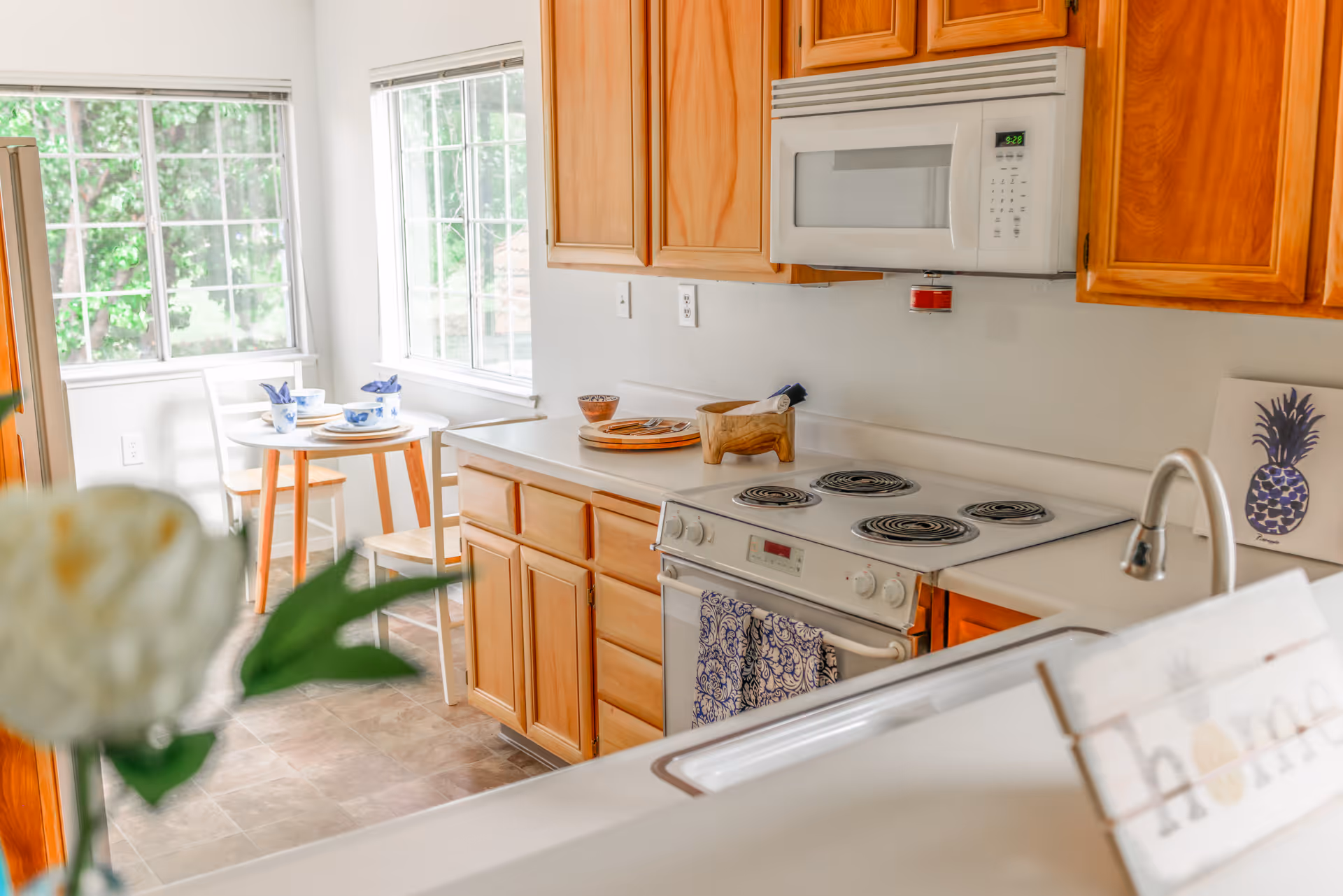Bright kitchen area with wooden cabinets, white electric stove and microwave, and a small dining table set near large windows letting in natural light. A flower and a decorative 'home' sign are visible in the foreground.
