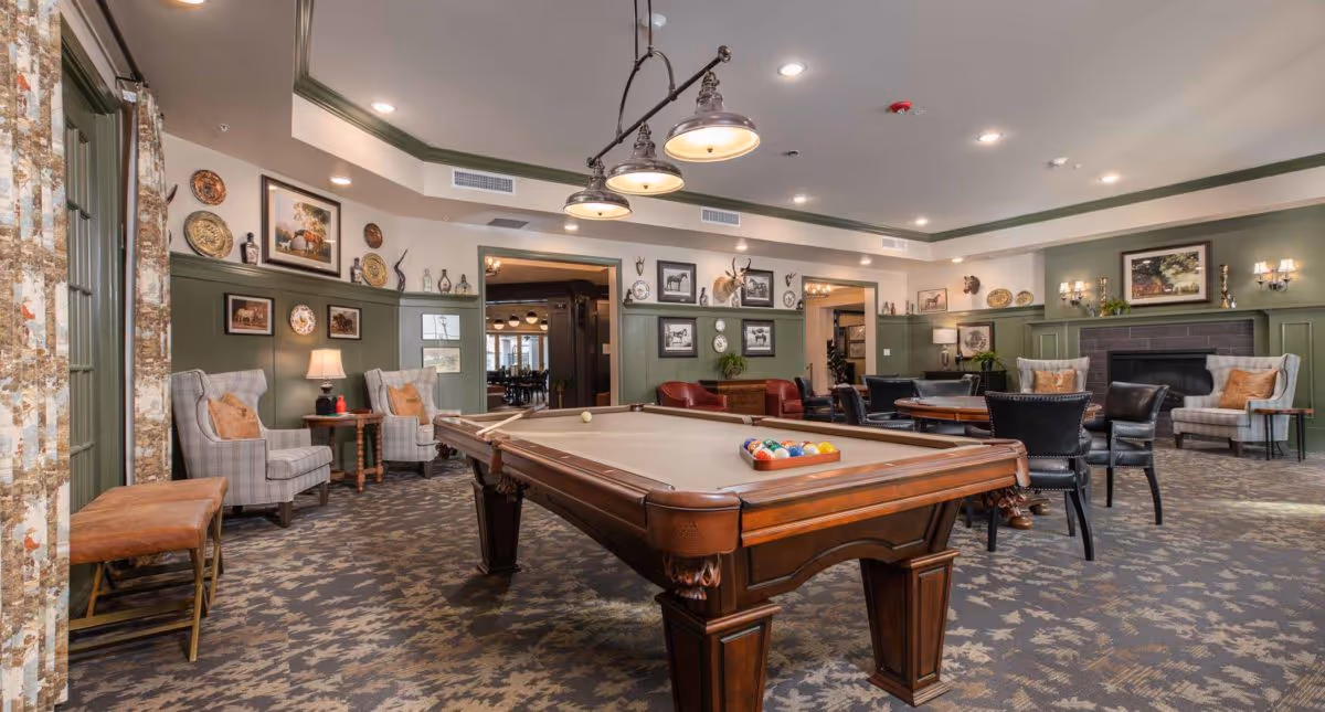 A spacious and well-lit common area featuring a pool table in the center, surrounded by comfortable seating including armchairs and leather chairs. The room has green and beige walls adorned with framed pictures, decorative plates, and mounted animal heads. There is a fireplace on one side and a patterned carpet covering the floor.