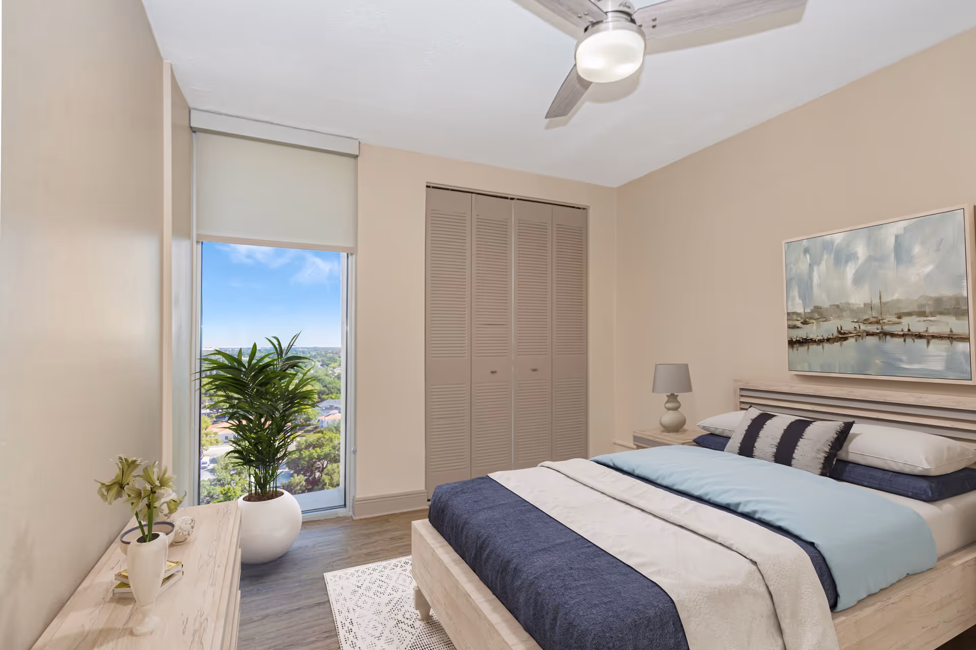 Bright furnished bedroom with a bed, bedside table and lamp, a large window with a potted plant, closet doors, and a ceiling fan.