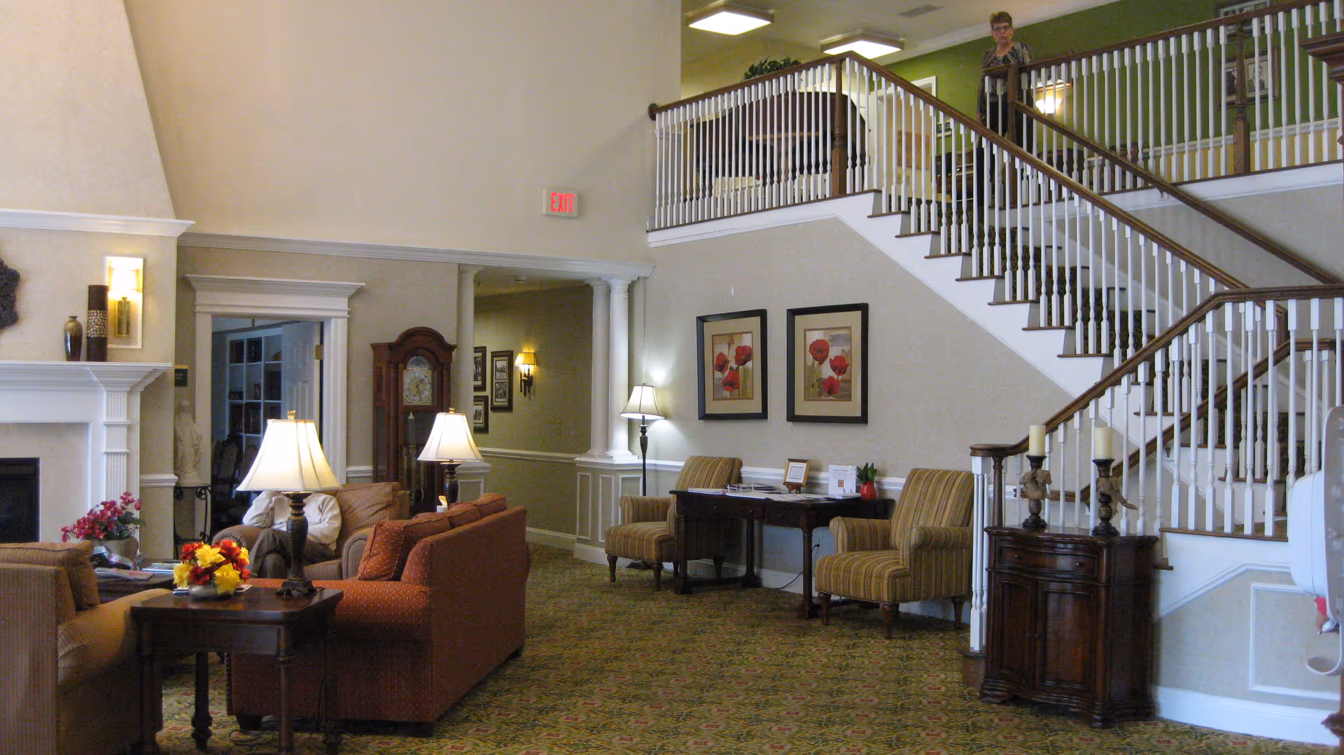 A spacious senior living center common area with comfortable seating including sofas and armchairs, a fireplace, table lamps, floral decorations, a grandfather clock, framed artwork on the walls, and a staircase with white spindles and wooden handrails. A person is standing on the upper level near the railing.