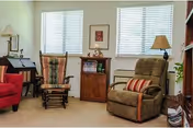 A cozy living room area with a red armchair, a wooden rocking chair with a cushion, a brown recliner with a striped pillow, a small wooden cabinet with decorative items, two windows with blinds, and two lamps on either side.
