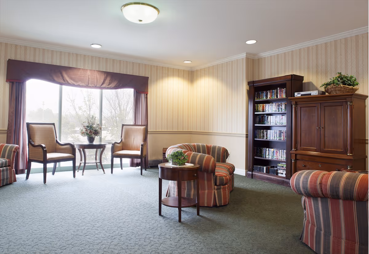 Comfortable sitting room with upholstered armchairs, small tables, a bookcase and a large window letting in daylight.