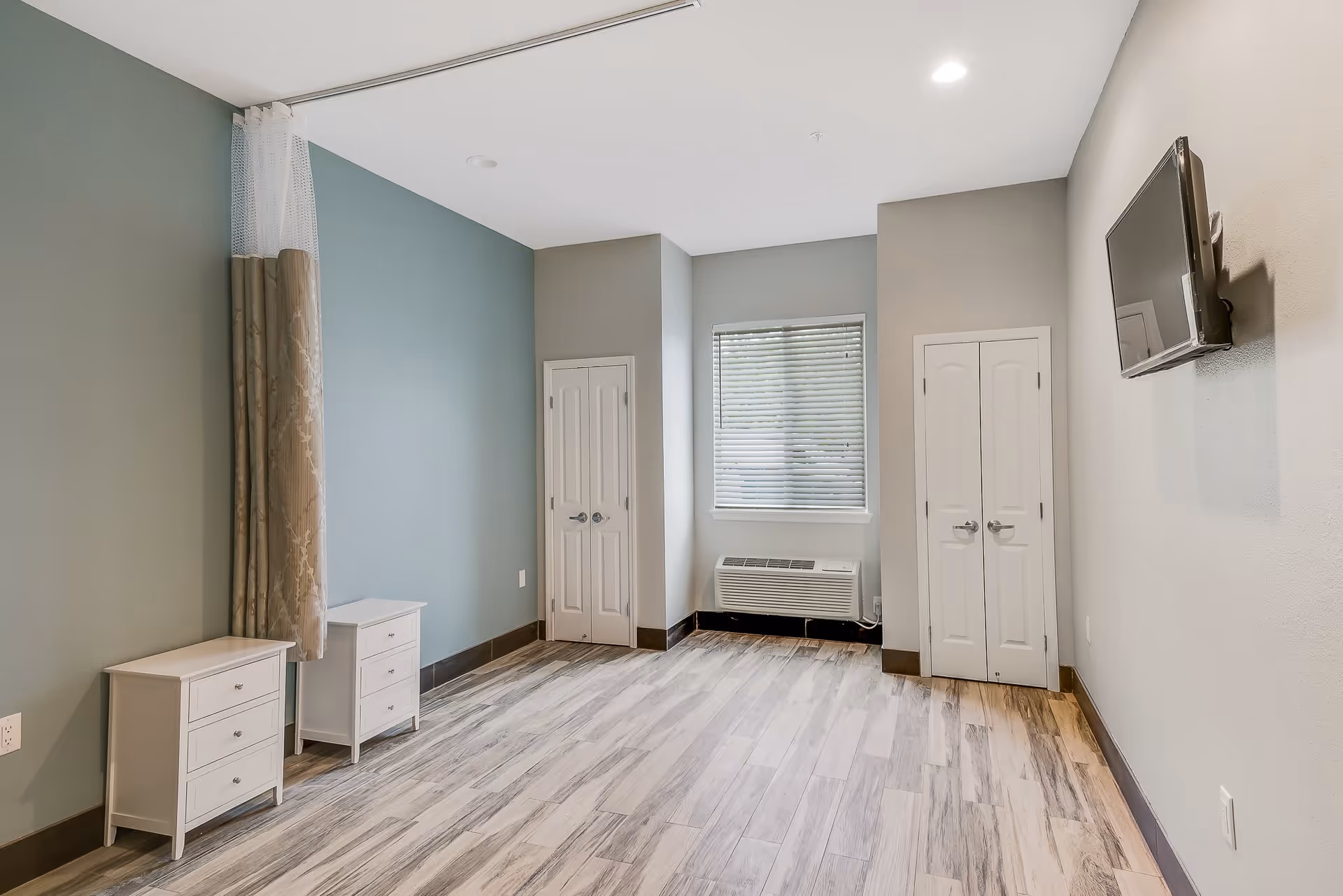Empty assisted-living bedroom with wood-look flooring, two small nightstands, a wall-mounted TV, window and closets.