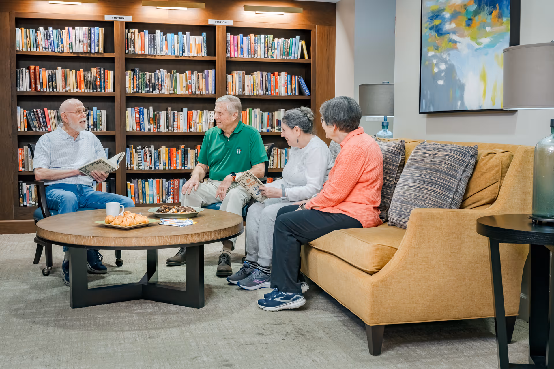 Four elderly people sitting and chatting in a cozy library room with bookshelves filled with books behind them. Two women and two men are seated around a round wooden coffee table with snacks and a cup on it. The room has a comfortable yellow couch, a side table with a lamp, and a colorful abstract painting on the wall.