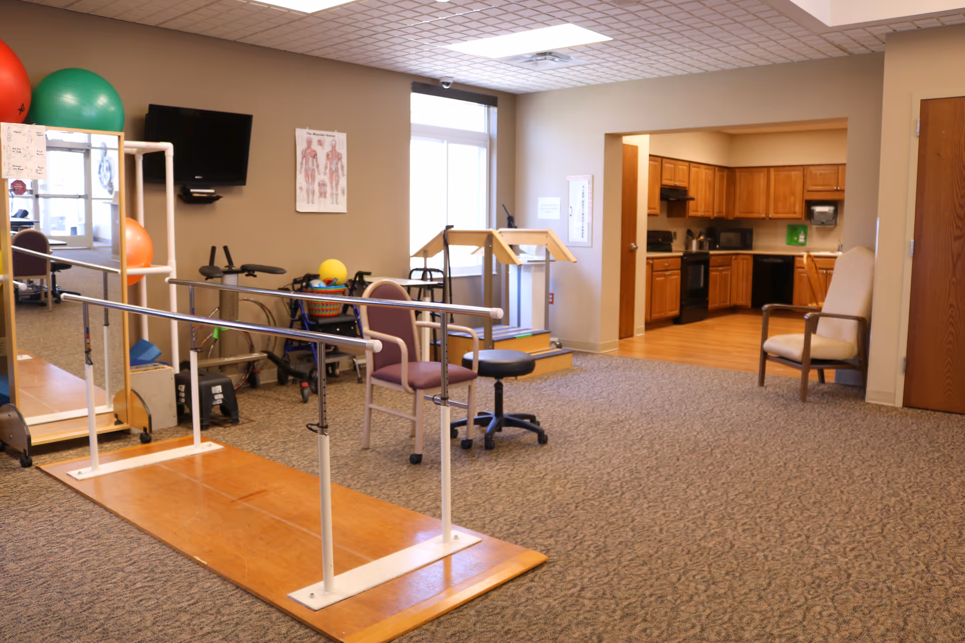 A therapy room with parallel bars for walking exercises, chairs, a stool, exercise balls, a walker, and a wall-mounted TV. There is a window letting in natural light and a doorway leading to a kitchen area with wooden cabinets and appliances.