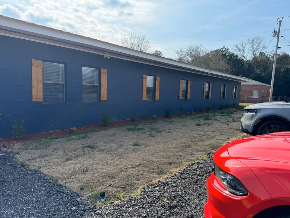Exterior view of a single-story building painted dark blue with wooden window shutters. There is a patch of grass and gravel in front of the building, and two parked cars, one red and one gray, are partially visible on the right side. Trees and a utility pole are in the background under a partly cloudy sky.