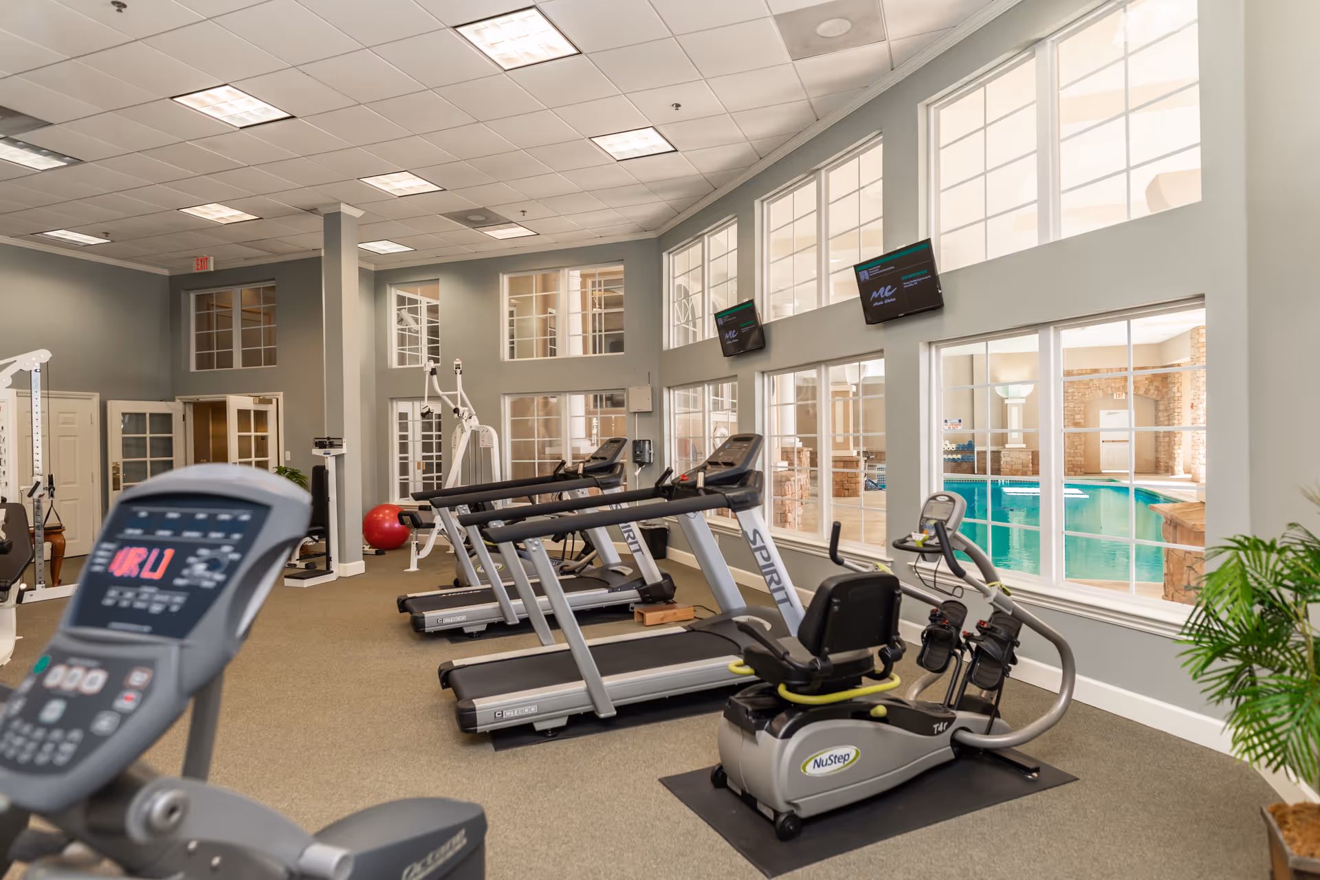 Bright fitness room with treadmills, exercise bikes and large windows overlooking an indoor pool.