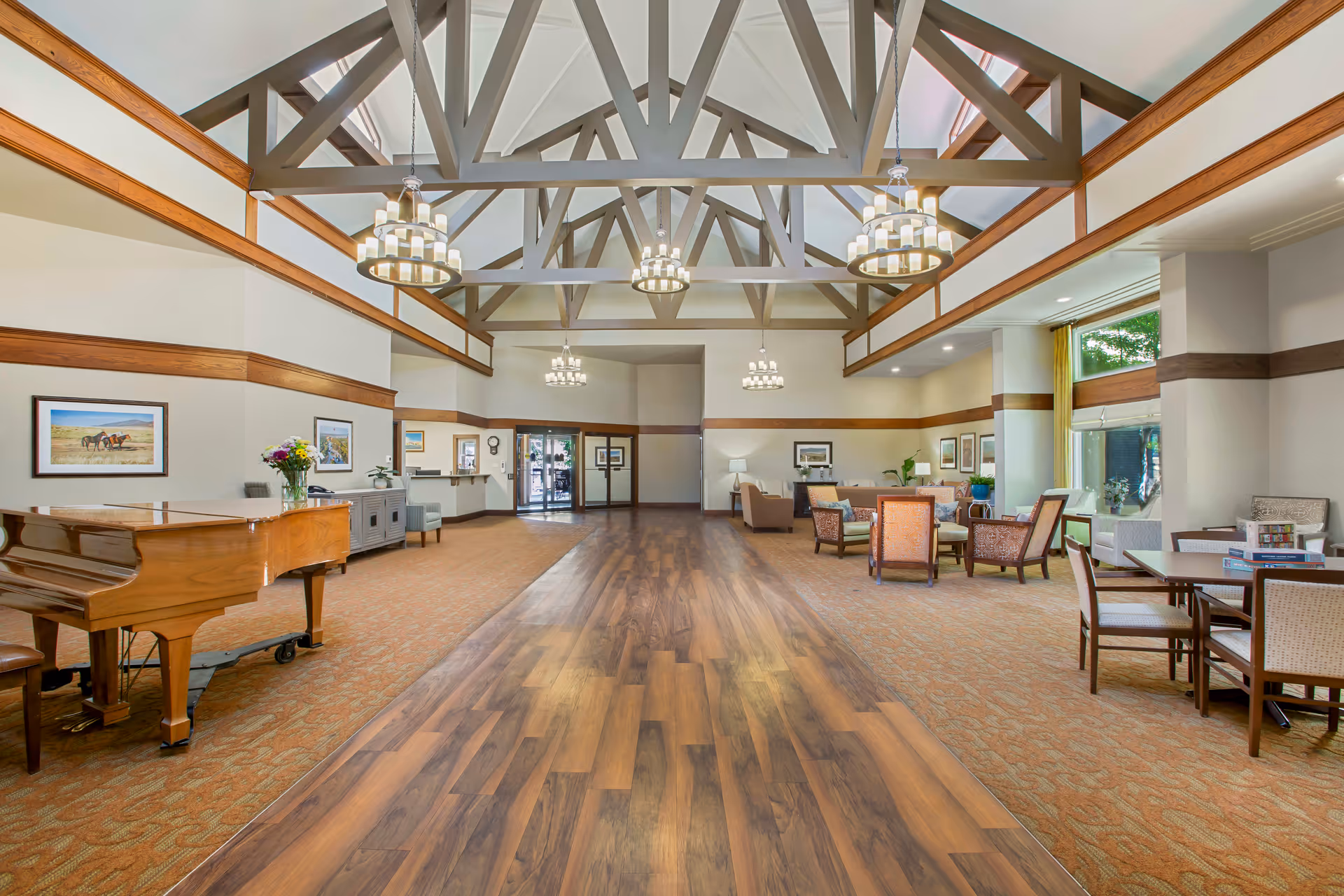 Spacious senior living facility common area with high vaulted ceiling featuring exposed wooden beams and multiple chandeliers. The room has a mix of wood and carpet flooring, a grand piano on the left, several seating areas with chairs and tables, large windows letting in natural light, and framed artwork on the walls.