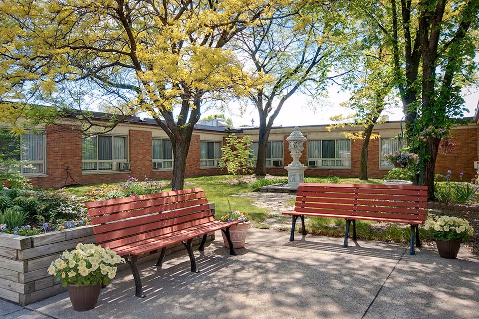 Sunny courtyard with two red benches, potted flowers, trees, and a low brick building in the background.
