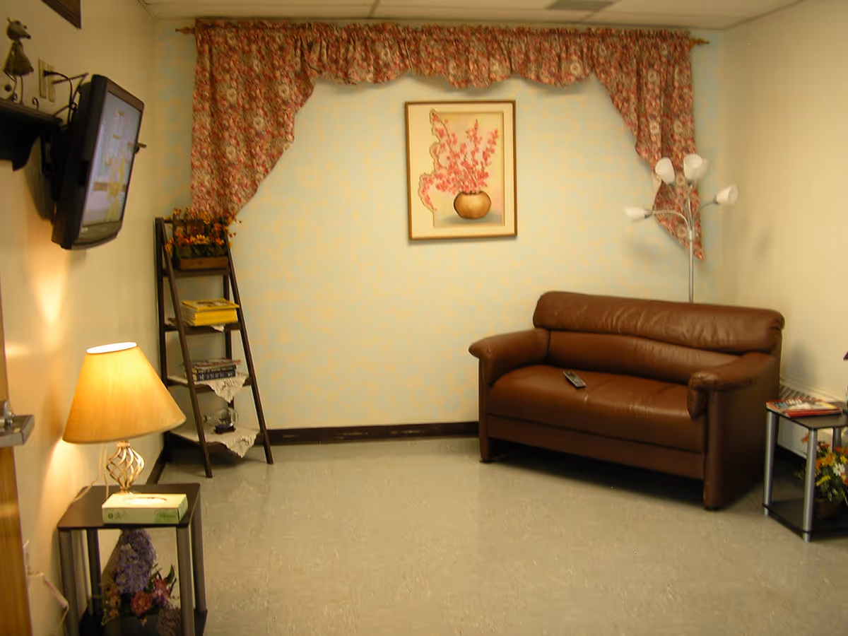 A small living room with a brown leather couch, a floor lamp with five white shades, a wall-mounted TV, a small side table with a lamp and tissue box, a ladder-style shelf with books and flowers, and a framed floral painting on a light blue wall with floral curtains.