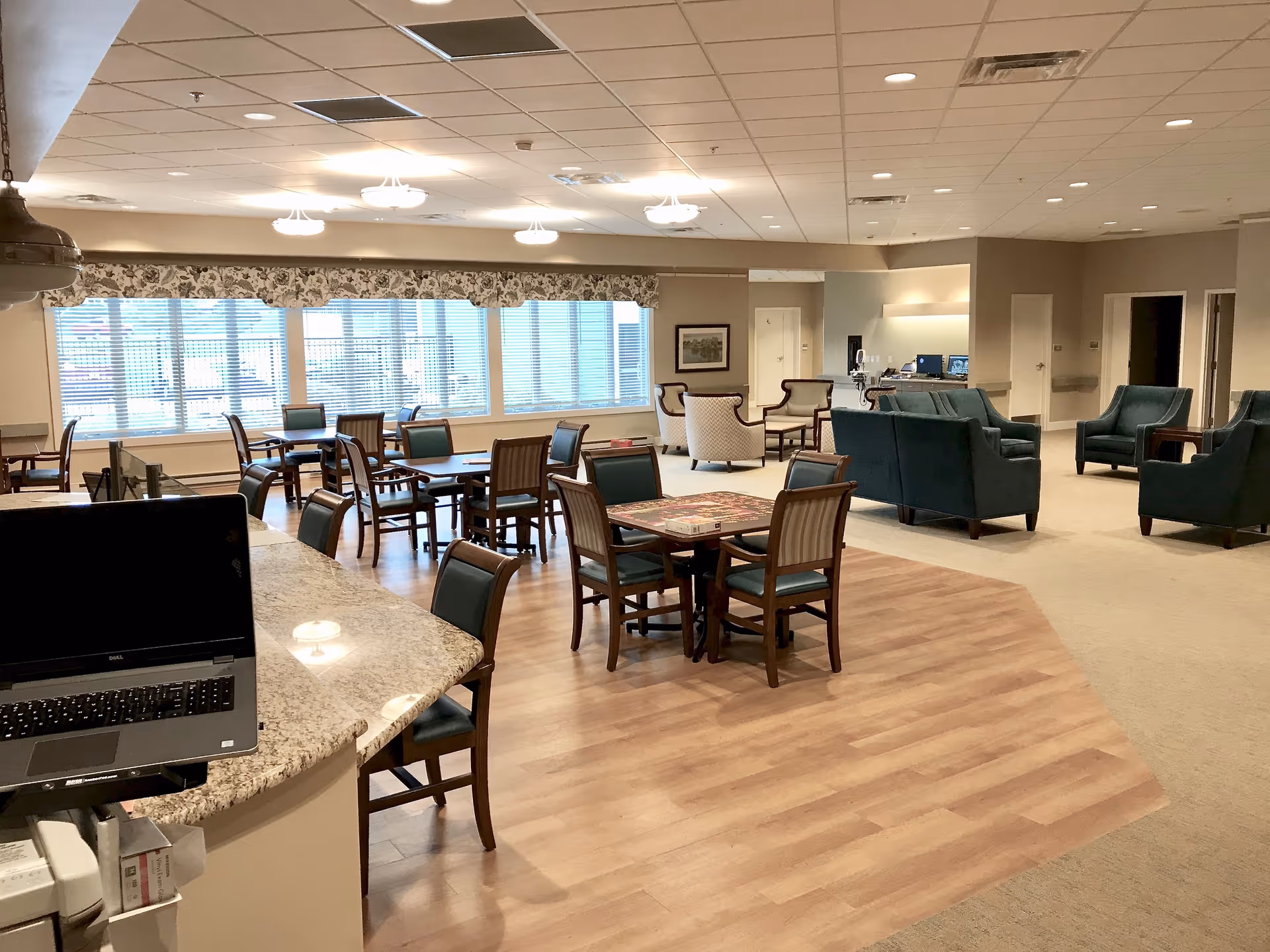 A spacious common area in a senior living facility featuring multiple tables and chairs arranged for dining or activities, a seating area with armchairs and sofas, large windows with blinds and floral valances allowing natural light, and a granite countertop with a laptop on it in the foreground.