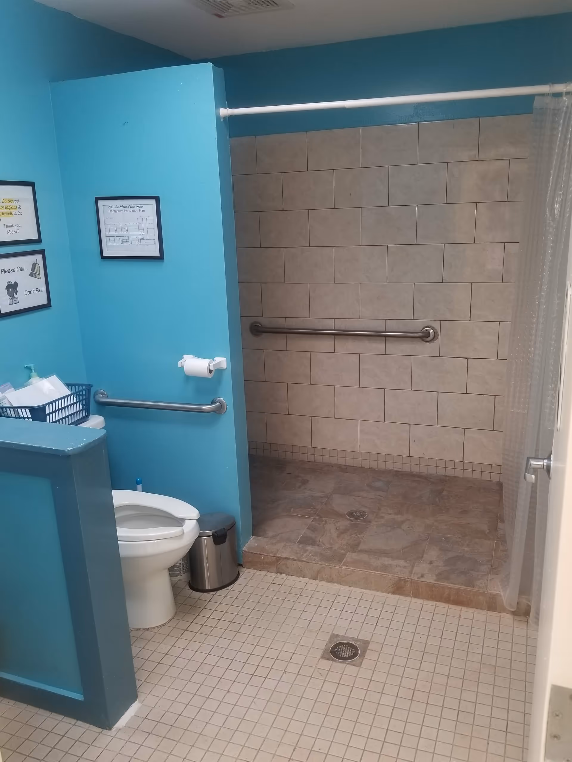 A bathroom with turquoise walls featuring a toilet with a grab bar beside it, a small trash can, and a walk-in tiled shower with a grab bar and a clear shower curtain.