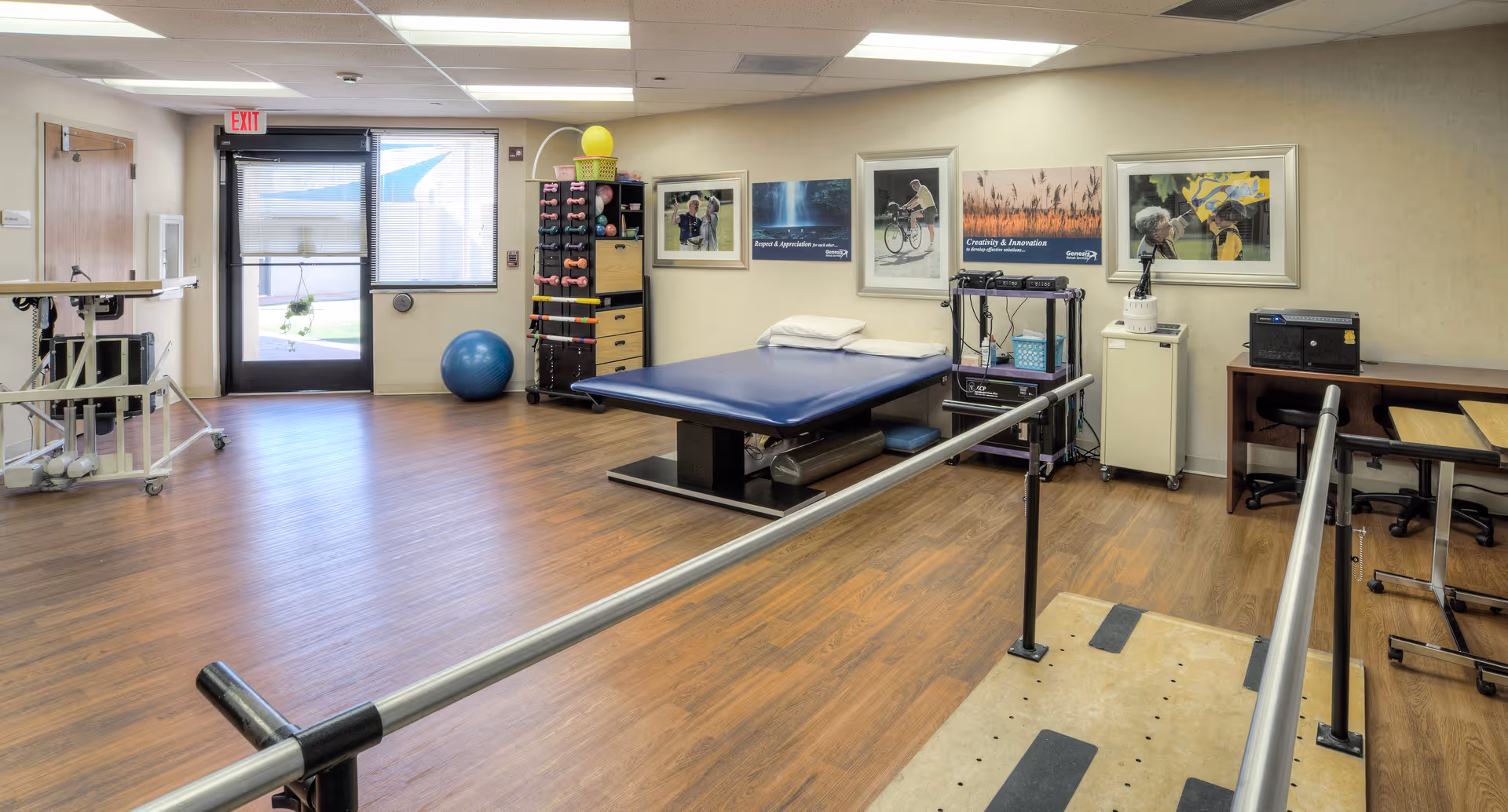 A rehabilitation therapy room with parallel bars for walking exercises, a padded treatment table with a pillow, exercise equipment including dumbbells and a large exercise ball, framed motivational posters on the wall, and a desk with a chair. The room has wood flooring and a door with a window leading outside.