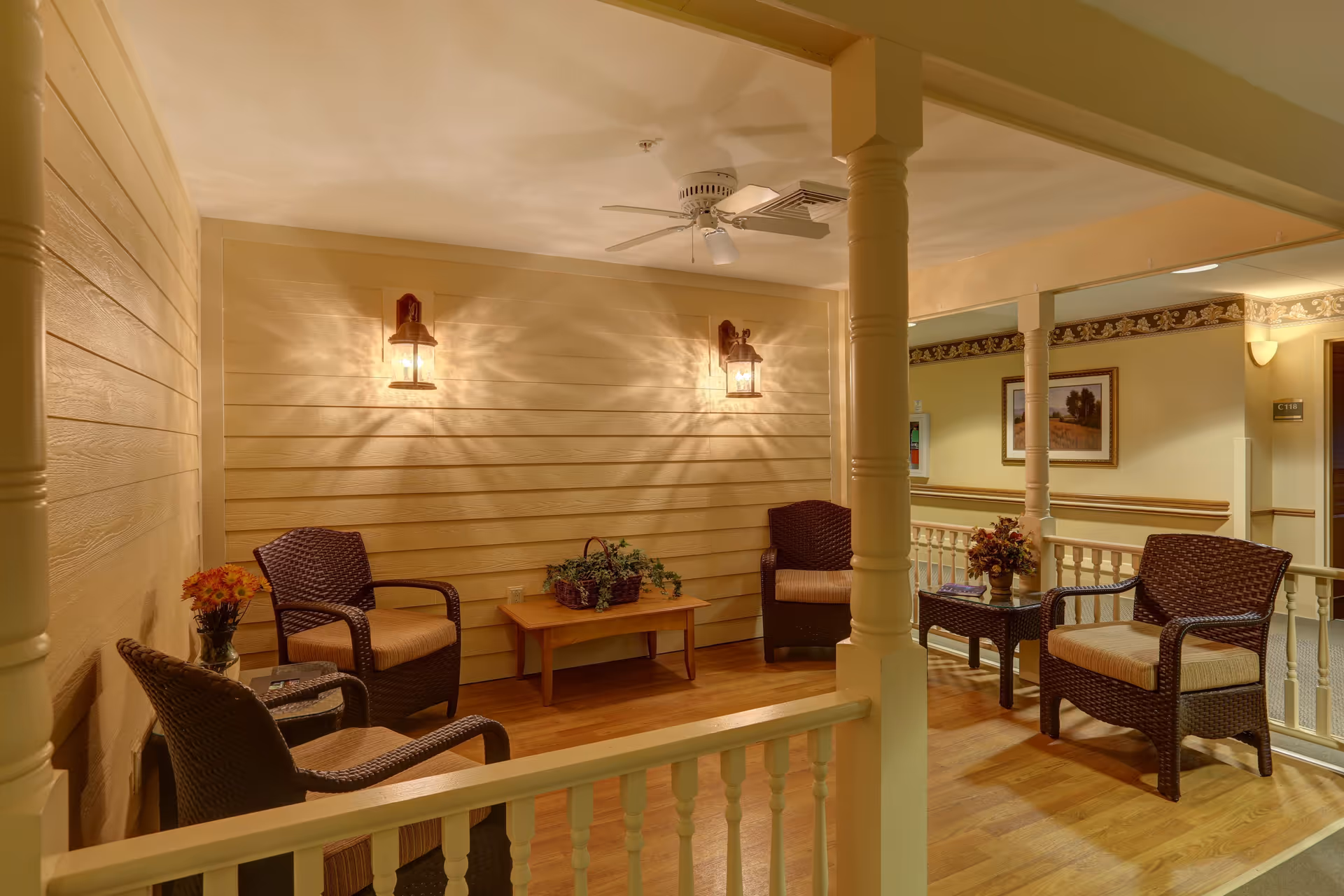 A cozy indoor seating area with four wicker chairs with cushions arranged around two small wooden tables. The walls are painted a warm beige with horizontal paneling, and two wall-mounted lantern-style lights illuminate the space. There are decorative plants on the tables, a ceiling fan above, and a railing separating the seating area from a hallway with framed artwork on the walls.