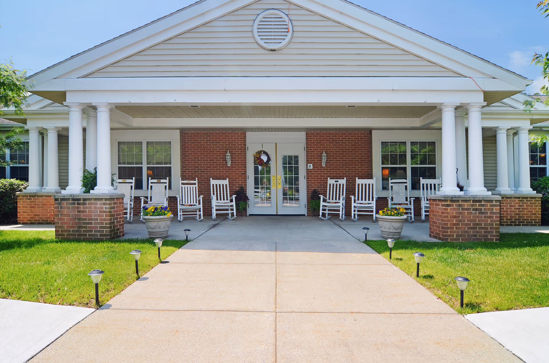 Front entrance of Celebration Villa of Lebanon PA featuring a covered porch with white columns, several white rocking chairs, potted flowers, and a concrete walkway leading to double glass doors with a wreath.