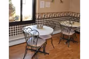 Small dining area with two round white tables and black metal chairs with cushions. The room has a checkered black and white wallpaper border and a large window letting in natural light. The floor is wooden.