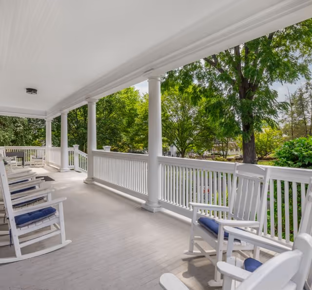 A covered white porch with several rocking chairs and a railing overlooking green trees.