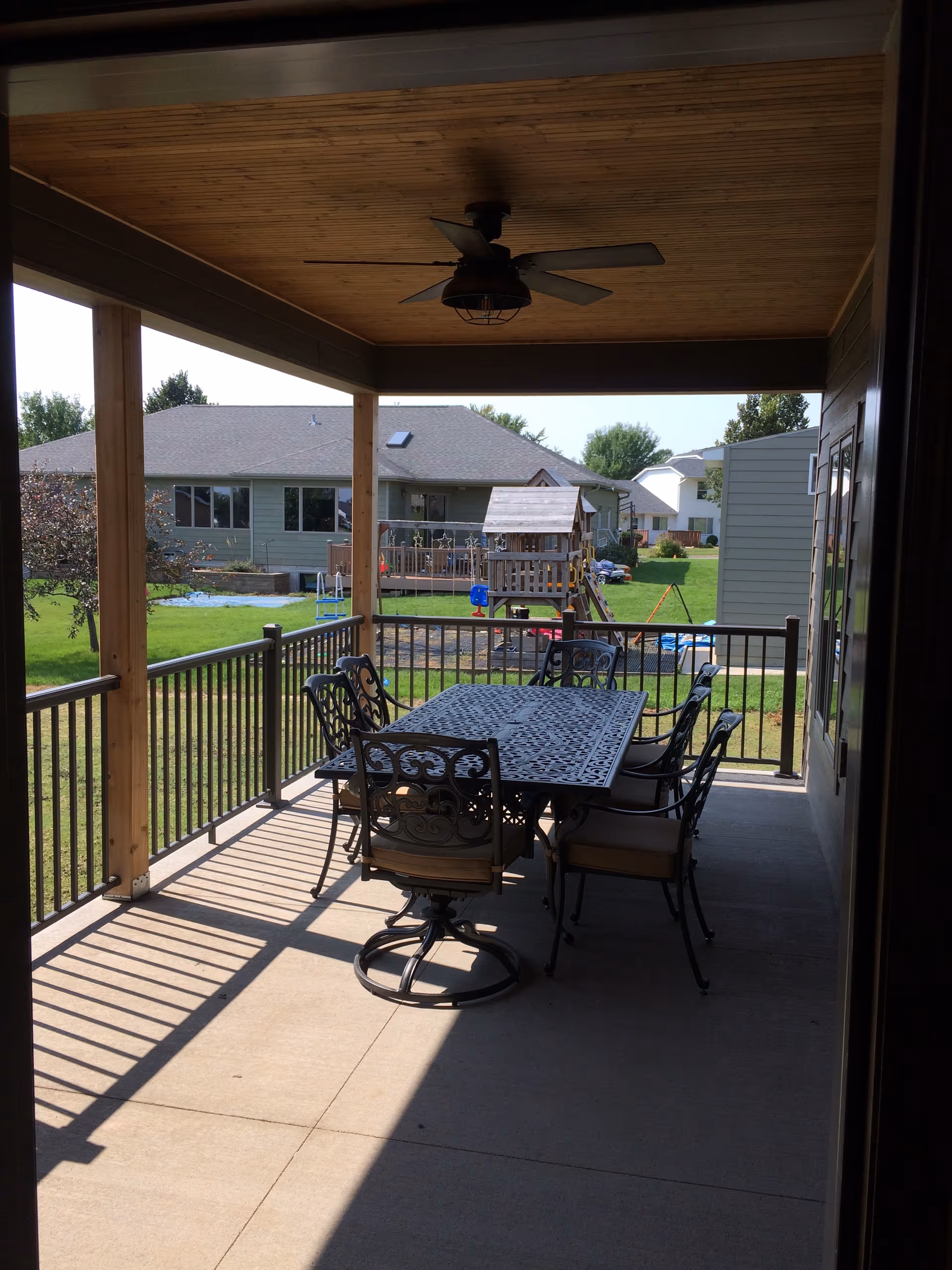 Covered backyard porch with a metal dining table and chairs under a ceiling fan, overlooking a lawn, neighboring houses, and a children's playset.