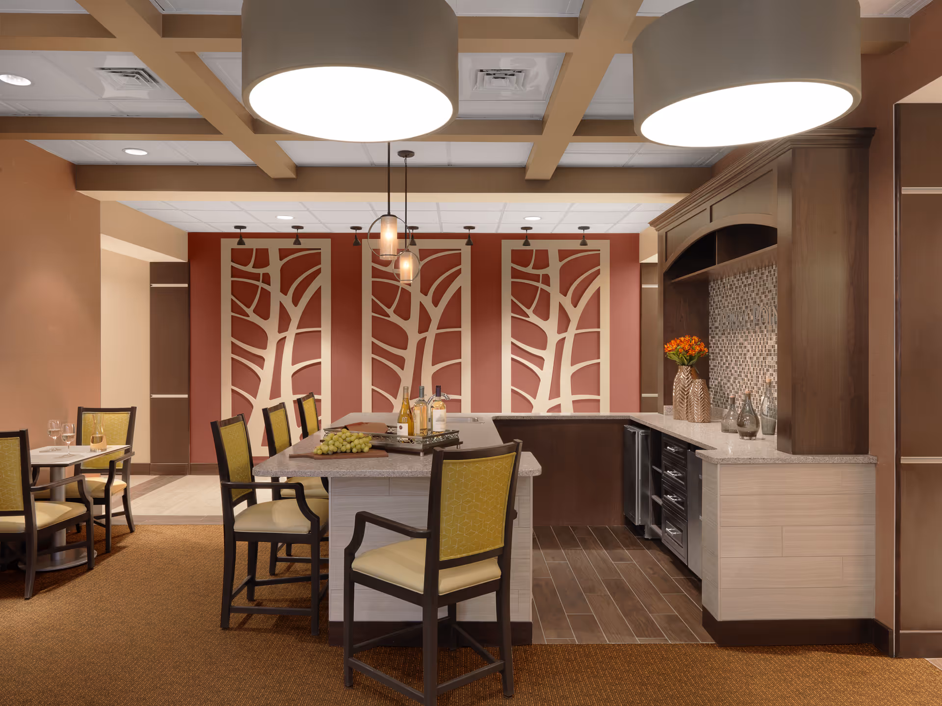 A modern dining and kitchen area in a senior living facility featuring a central island with chairs, bottles of wine, and grapes on the counter. The background has decorative wall panels with abstract tree designs, pendant lights hanging from the ceiling, and a countertop with vases and flowers. The space has warm tones with brown, beige, and red accents.