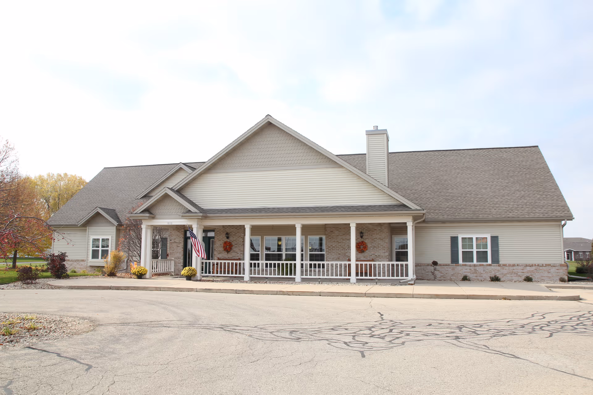 Single-story tan and brick senior living building with a covered front porch, American flag, and paved driveway in front.