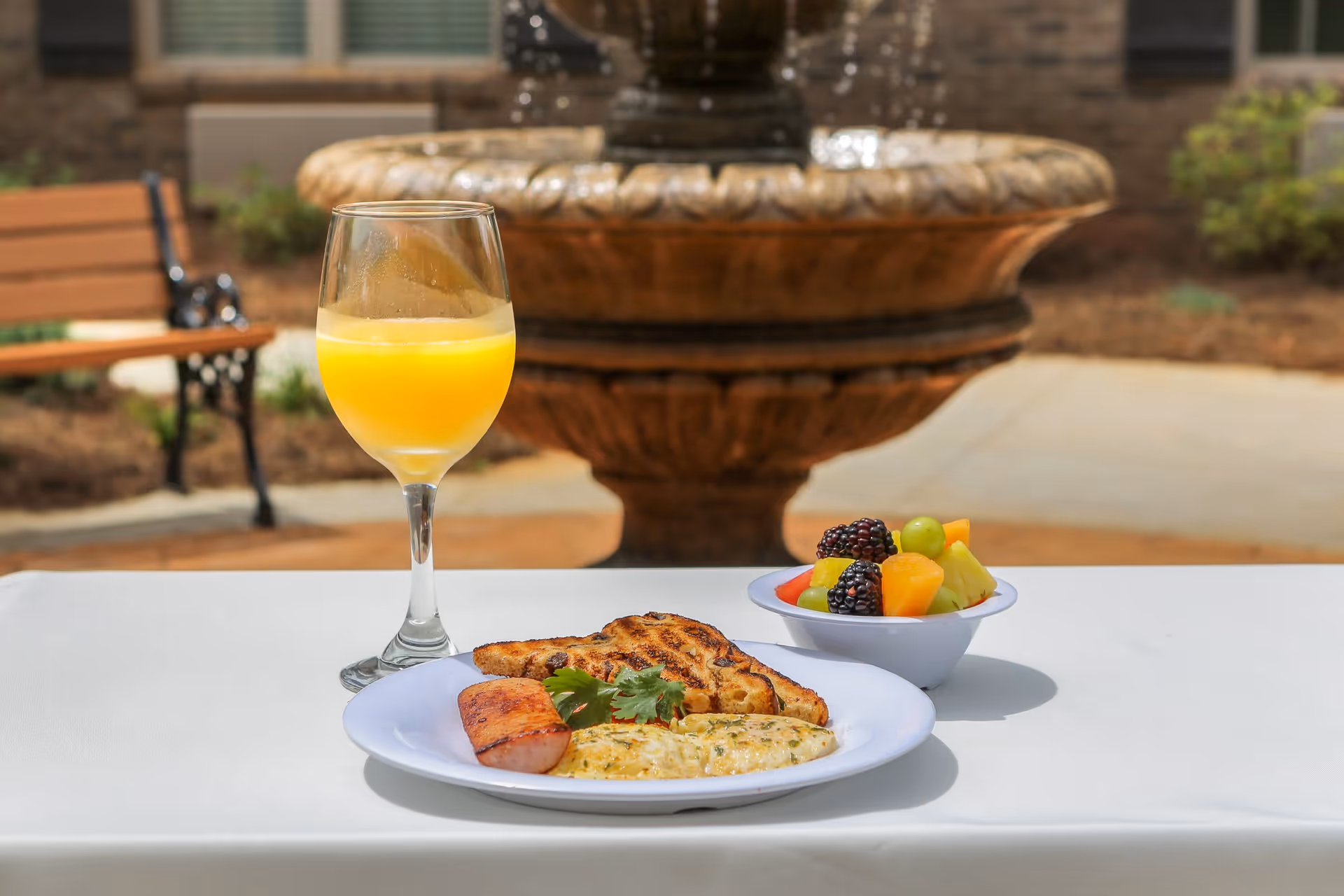 A breakfast plate with omelette, grilled toast and sausage, a bowl of fruit and a glass of orange juice on a table outdoors in front of a fountain.