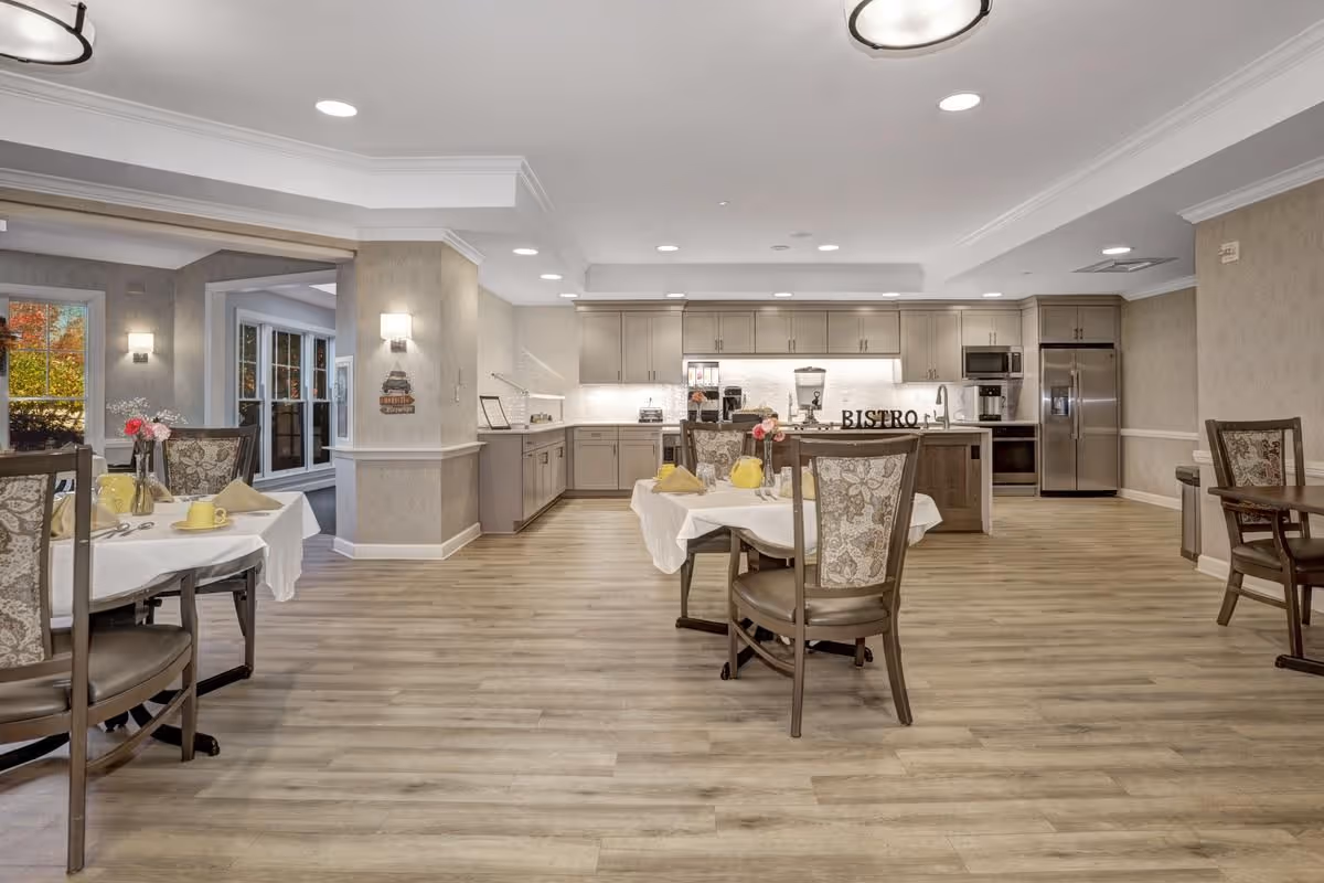A spacious dining area in a senior living facility with several tables covered in white tablecloths, each set with yellow cups, napkins, and small flower arrangements. The room features light wood flooring, beige walls, and a modern kitchen area in the background with gray cabinetry, stainless steel appliances, and a sign that reads 'BISTRO'. Large windows and soft lighting create a warm and inviting atmosphere.