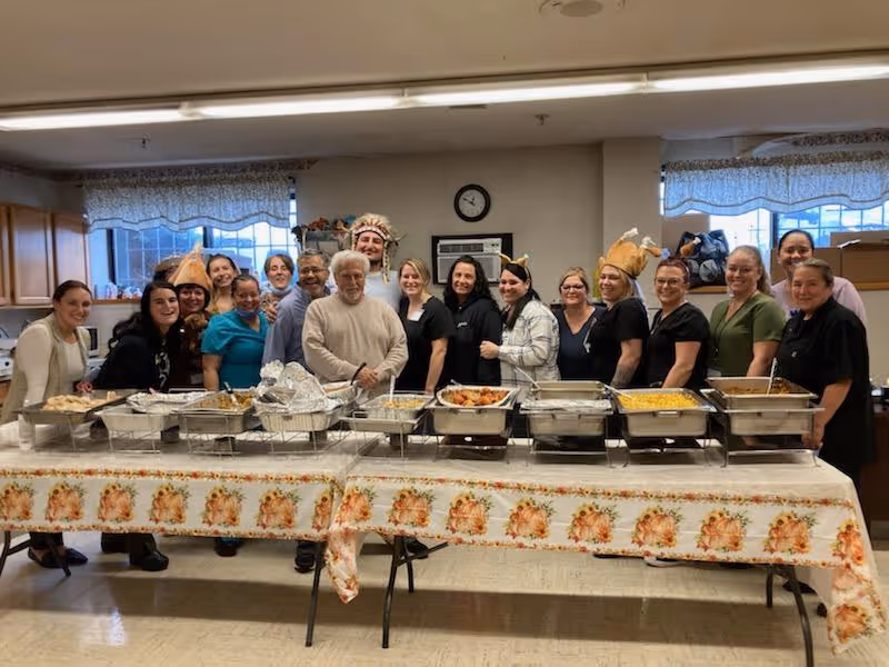 A group of people standing behind a long table covered with a floral tablecloth and trays of food in a kitchen or dining area. The group is smiling and some are wearing festive hats. The room has windows with lace curtains and wooden cabinets.