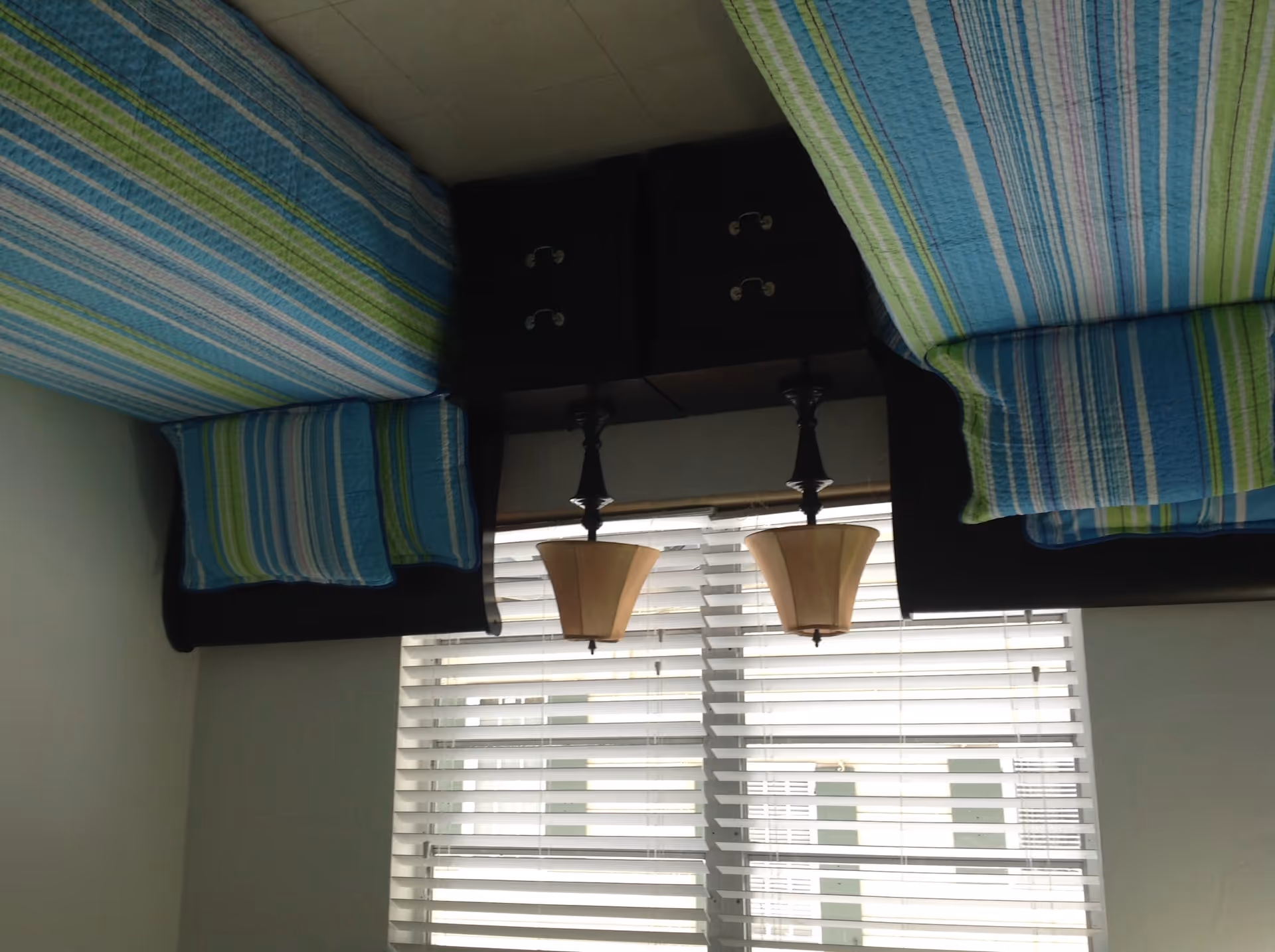 A bedroom with two twin beds featuring blue, green, and white striped bedding and matching pillows. Between the beds is a dark wooden nightstand with two drawers and two lamps with beige lampshades. Behind the nightstand is a window with white blinds.