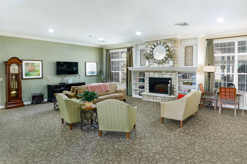 A spacious living room in an assisted living facility featuring a stone fireplace with a round mirror above it, built-in shelves with books and decor, a beige sofa with a red and white throw, two green patterned armchairs around a small round table with a plant, a beige loveseat, a wooden grandfather clock, a flat-screen TV mounted on a green wall, framed artwork, and large windows with curtains allowing natural light.