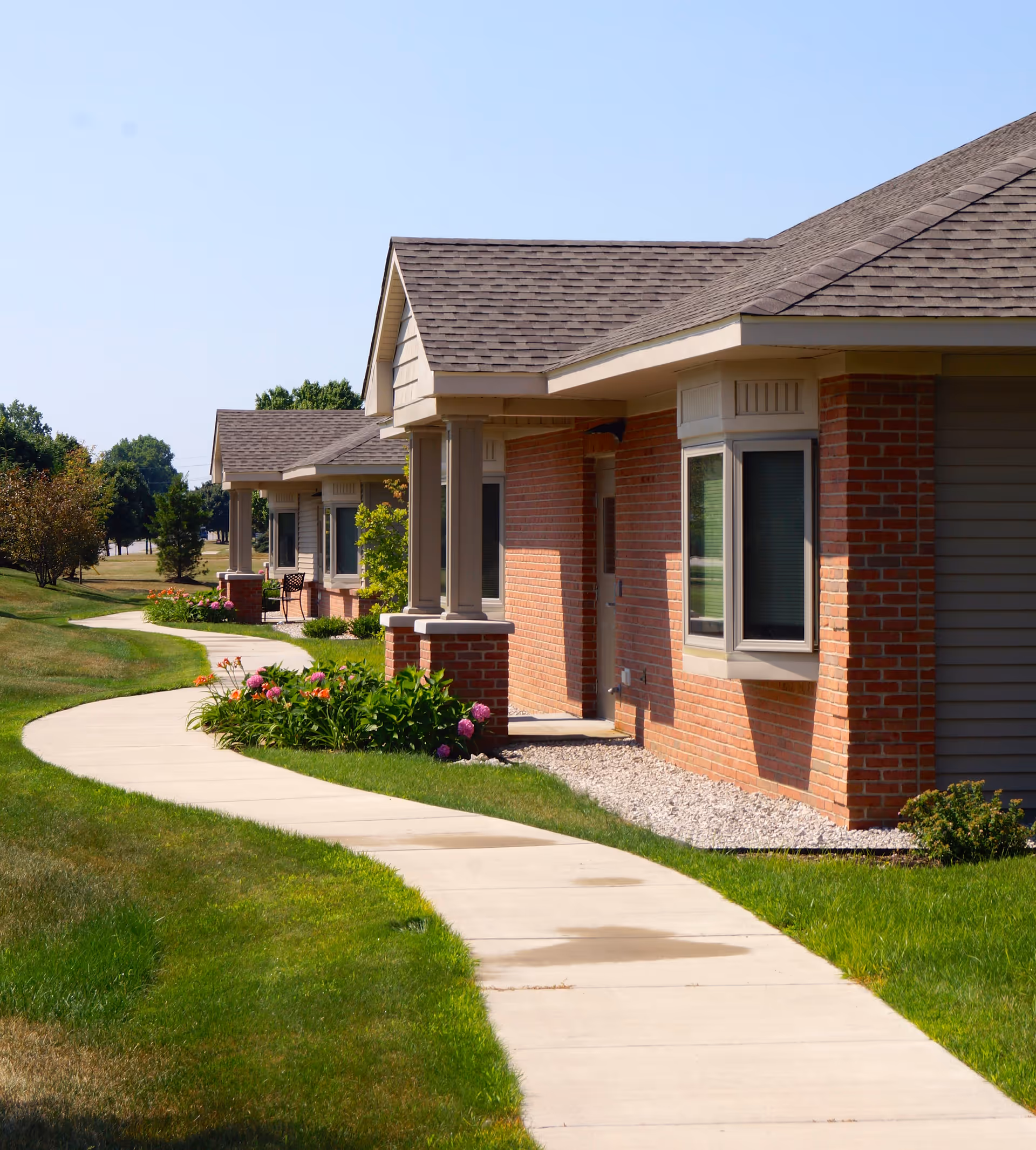 A curved concrete walkway bordered by green grass and flower beds leads past a row of single-story brick and siding buildings with gabled roofs under a clear blue sky.