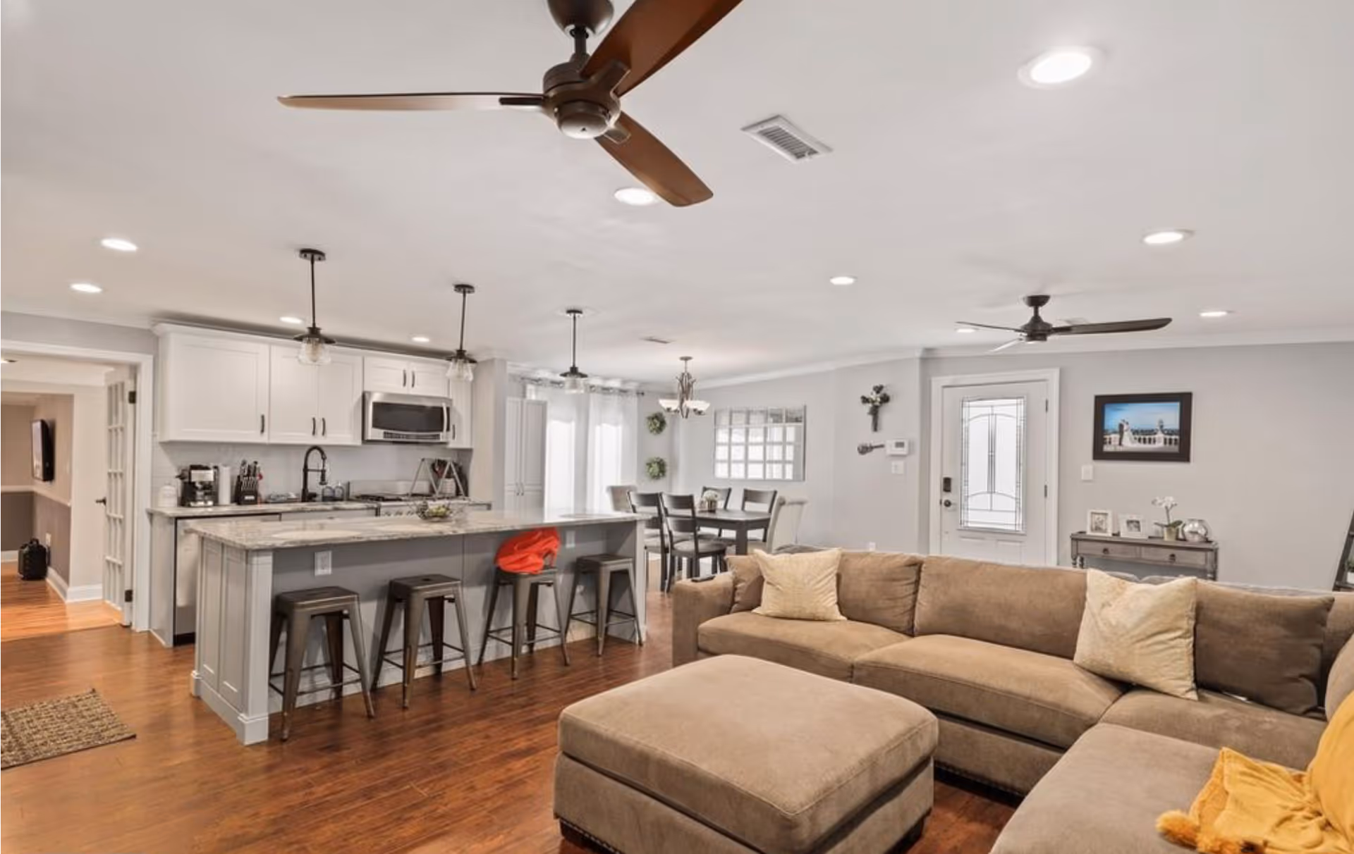 Open concept living area featuring a large beige sectional sofa with pillows and an ottoman, a kitchen with white cabinets, a marble countertop island with four metal bar stools, and a dining area with a table and chairs. The room has wooden flooring, ceiling fans, recessed lighting, and a glass-paneled door leading outside.