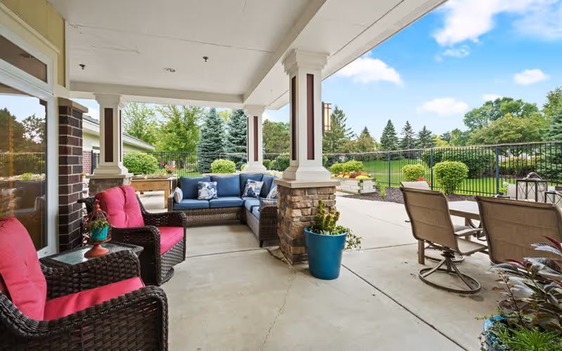 Covered outdoor patio area with wicker furniture including two chairs with red cushions and a sectional sofa with blue cushions. There is a small table with a plant, a blue planter with greenery, and a dining table with four chairs. The patio overlooks a green lawn with trees and shrubs under a partly cloudy sky.
