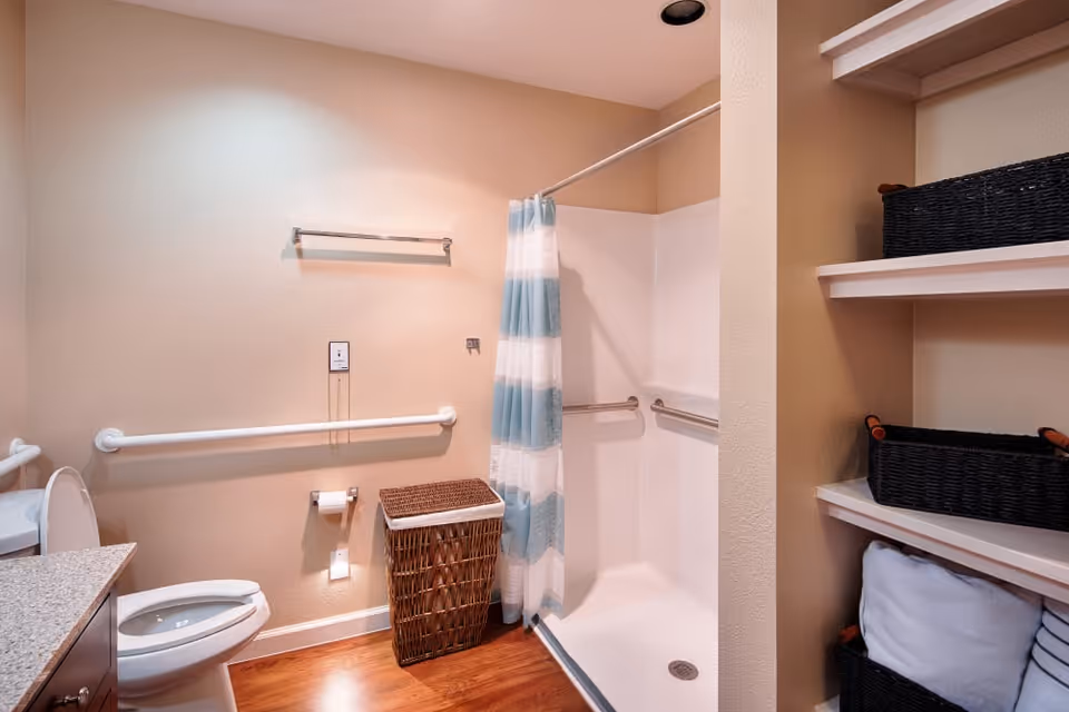 Accessible bathroom with a toilet, grab bars, a walk-in shower with a striped curtain, a wicker laundry basket, and shelving with baskets and towels.