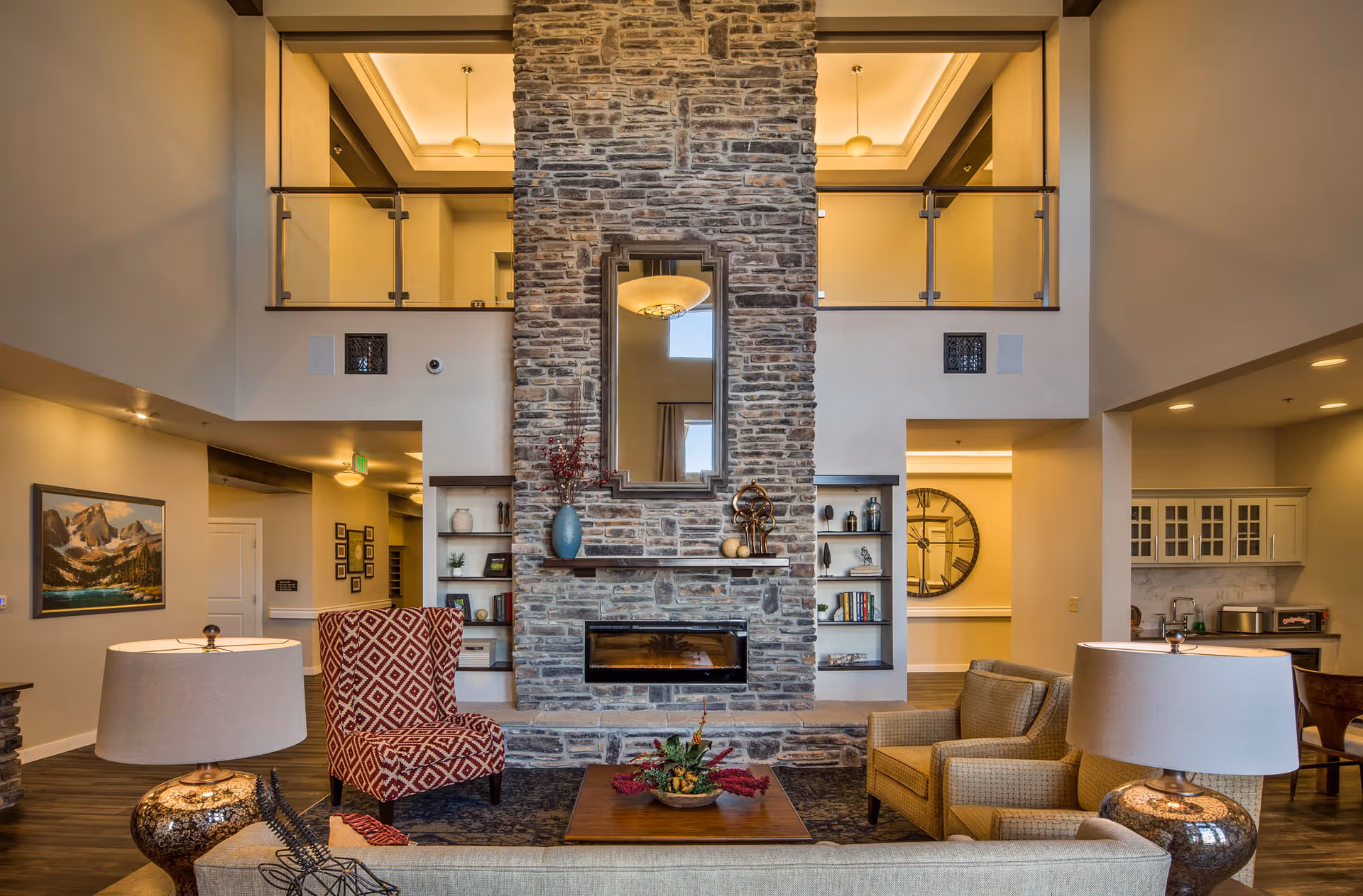Spacious two-story communal living room with a central stone fireplace, seating areas, and an upper balcony overlook.