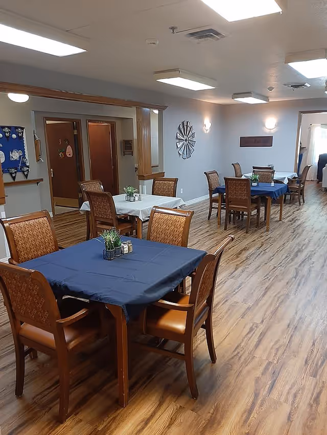 A dining area in a senior living facility with several wooden tables covered with blue and white tablecloths, each surrounded by four wooden chairs with cushioned seats. The room has wood-patterned flooring, light gray walls, ceiling lights, and decorative wall art. There are doorways and a bulletin board visible in the background.