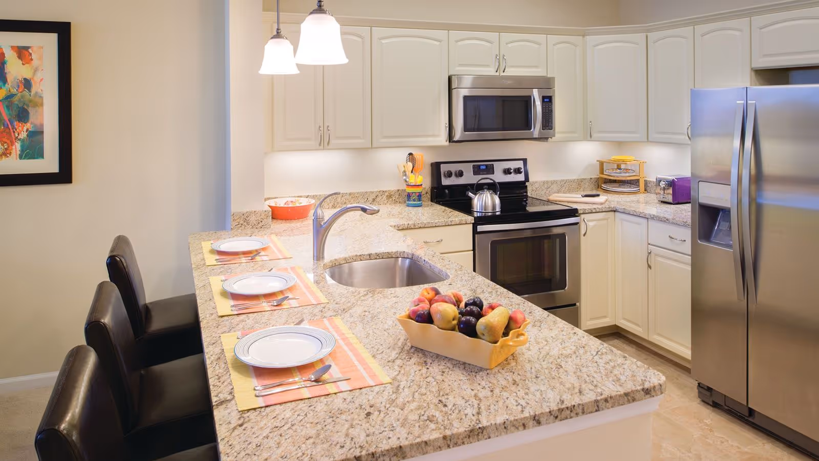Modern kitchen with granite countertops, a sink, stainless steel appliances including a refrigerator, oven, and microwave. Three place settings with plates, forks, and spoons are arranged on the counter with black cushioned chairs. A bowl of assorted fruit is placed on the counter, and a colorful abstract painting hangs on the wall.