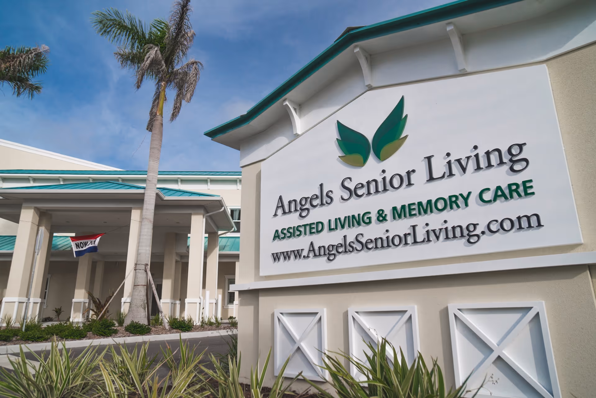 Exterior view of Angels Senior Living at Sarasota facility showing the building entrance with a covered driveway, palm trees, and a large sign displaying the facility name, services offered (Assisted Living & Memory Care), and website URL.