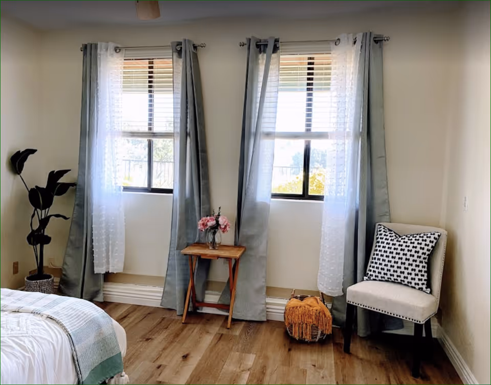 A cozy bedroom corner with two windows covered by sheer white and gray curtains. Below the windows is a small wooden table with a vase of pink flowers. To the right is a beige upholstered chair with a black and white patterned pillow. A woven basket with a fringed blanket is on the floor next to the chair. Part of a bed with white bedding and a green throw is visible on the left side. The room has light-colored walls and wooden flooring.